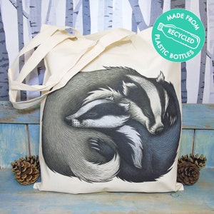 Könnte beinhalten: Eine weiße Einkaufstasche mit einer Schwarzweißillustration von zwei Dachsen, die sich aneinanderkuscheln. Die Tasche hat einen grünen Kreis mit dem Text "Hergestellt aus recycelten Plastikflaschen".
