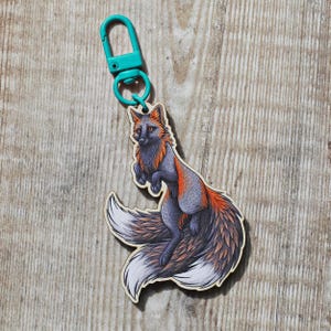 May include: A keyring featuring a fox illustration with grey and orange fur and a large white-tipped tail. The fox is attached to a bright teal plastic lobster clasp. The design is printed on a flat, light-coloured material with a visible wood grain.