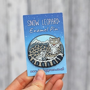 以下が含まれることがあります： 雪ヒョウが安らかに休んでいるエナメルピン。ピンは「SNOW LEOPARD Enamel Pin」の文字が入った青色の背景にセットされています。雪ヒョウは斑点のあるコートを着て、丸まっています。