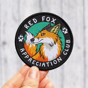 May include: A circular embroidered patch featuring a red fox illustration against a teal background. The black border contains the white text RED FOX APPRECIATION CLUB and two white paw prints. The fox has orange, white, and black fur details.