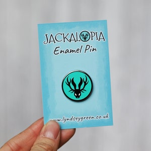 Op de afbeelding: Een turquoise emaille pin met een zwart silhouet van een jackalope, een mythisch wezen met gewei en konijnenoren. De pin heeft de tekst "Jackalopia Enamel Pin" en "www.lyndseygreen.co.uk".