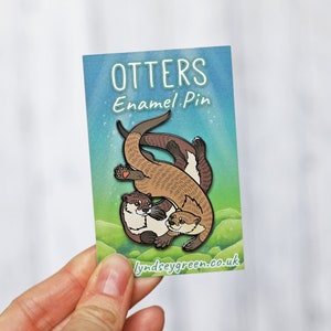 以下が含まれることがあります： 2匹のラッコが泳いでいる茶色と黒のエナメルピン。ピンは「Otters Enamel Pin」と「lyndseygreen.co.uk」のテキストが書かれたカードの上にあります。