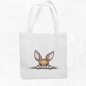 May include: White canvas tote bag with a brown rabbit peeking out from behind the top edge. The rabbit has brown fur, long ears, and pink nose.