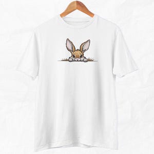 May include: White t-shirt with a brown rabbit peeking out from behind a green bush. The rabbit has brown eyes and long ears.
