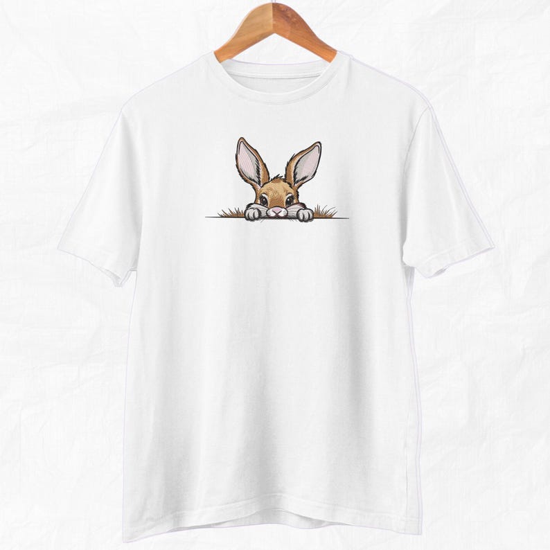May include: White t-shirt with a brown rabbit peeking out from behind a green bush. The rabbit has brown eyes and long ears.