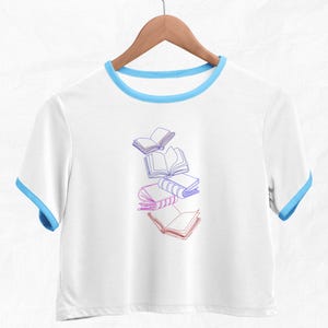 May include: White cropped t-shirt with a blue ringer neckline and a graphic of five books in a gradient of pink and purple.