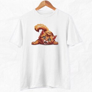 Op de afbeelding: Wit t-shirt met een cartoon kat illustratie. De kat is oranje en bruin met grote ogen en een speelse pose. Het t-shirt hangt aan een houten hanger. Het ontwerp is gecentreerd.