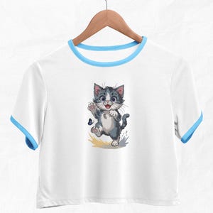 May include: White crop top with blue trim featuring a cartoon kitten illustration. The kitten is grey and white with blue eyes, waving at a blue butterfly. The shirt is hanging on a wooden hanger.
