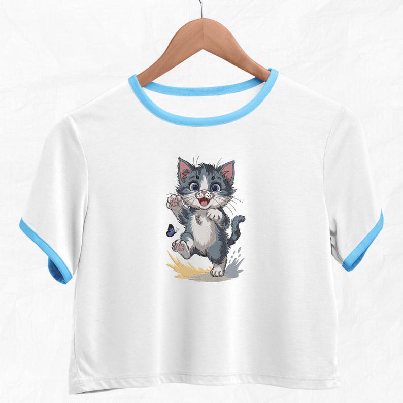 May include: White crop top with blue trim featuring a cartoon kitten illustration. The kitten is grey and white with blue eyes, waving at a blue butterfly. The shirt is hanging on a wooden hanger.