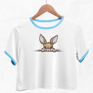May include: White t-shirt with a blue trim around the neckline. A cartoon bunny rabbit is peeking out from behind a patch of grass on the front of the shirt.