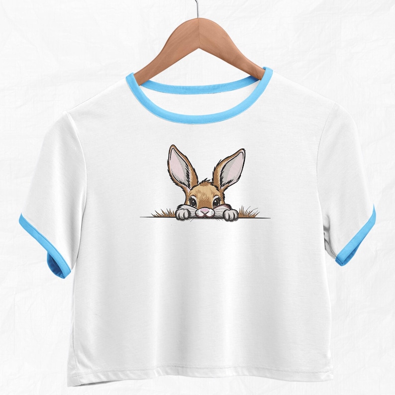 May include: White t-shirt with a blue trim around the neckline. A cartoon bunny rabbit is peeking out from behind a patch of grass on the front of the shirt.