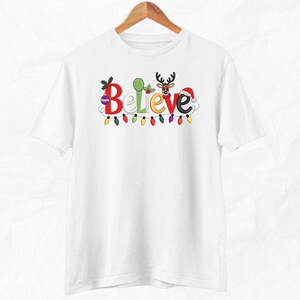 May include: White t-shirt with the word "Believe" in colorful, festive lettering. The "B" is decorated with holly, the "e" with a reindeer, and the "e" with a Santa hat. A string of colorful Christmas lights runs below the word.