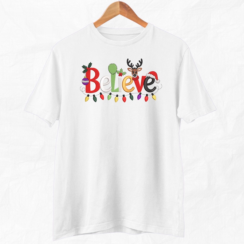May include: White t-shirt with the word "Believe" in colorful, festive lettering. The "B" is decorated with holly, the "e" with a reindeer, and the "e" with a Santa hat. A string of colorful Christmas lights runs below the word.