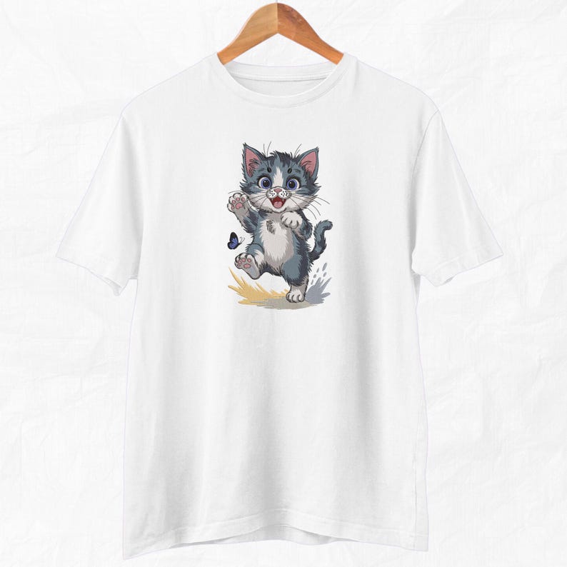 May include: White t-shirt with a cartoon kitten design. The kitten has grey and white fur, a blue butterfly, and a happy expression. The cat is mid-step, with one paw raised. The design is printed on a white t-shirt, suitable for cat enthusiasts.
