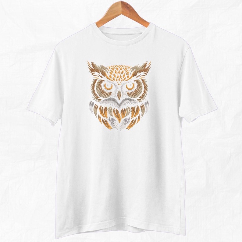 Op de afbeelding: Wit T-shirt met een afbeelding van een uil met bruine en gouden veren. De ogen van de uil zijn geel.