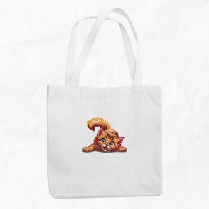 Op de afbeelding: Witte draagtas met een illustratie van een oranje tabby kat. De kat staat in een speelse pose, met zijn staart omhoog. De tas heeft twee witte handvatten en is gemaakt van een effen witte stof. De kat is oranje en bruin.