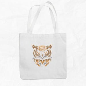 Op de afbeelding: Witte tote bag met een bruin en gouden uil ontwerp. De ogen van de uil zijn groot en zwart, en zijn veren zijn gedetailleerd.