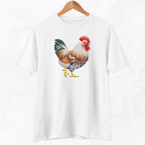 May include: White t-shirt with a colorful rooster illustration. The rooster has a red comb and wattle, brown and white feathers, and blue and white markings.