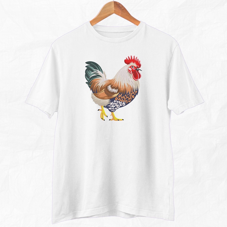 May include: White t-shirt with a colorful rooster illustration. The rooster has a red comb and wattle, brown and white feathers, and blue and white markings.