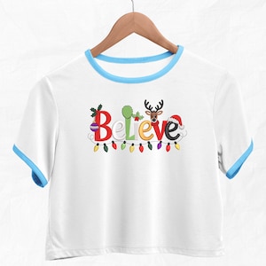 May include: White crop top with blue trim and sleeves. The word "Believe" is embroidered in colorful letters, with Christmas-themed embellishments. A string of colorful lights is below the word. The shirt is hanging on a wooden hanger.