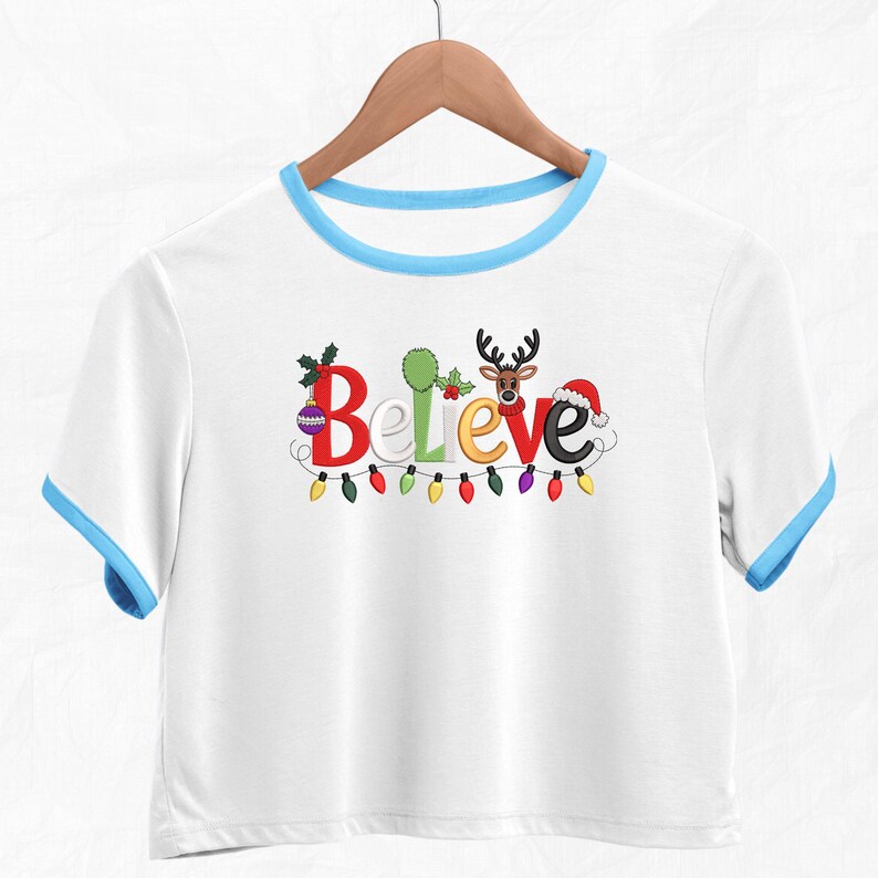 May include: White crop top with blue trim and sleeves. The word "Believe" is embroidered in colorful letters, with Christmas-themed embellishments. A string of colorful lights is below the word. The shirt is hanging on a wooden hanger.