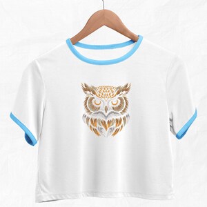 Op de afbeelding: Wit gecropped T-shirt met een blauwe rand rond de halslijn en mouwen. Het shirt heeft een afbeelding van een gestileerde uil met bruine en gouden veren.
