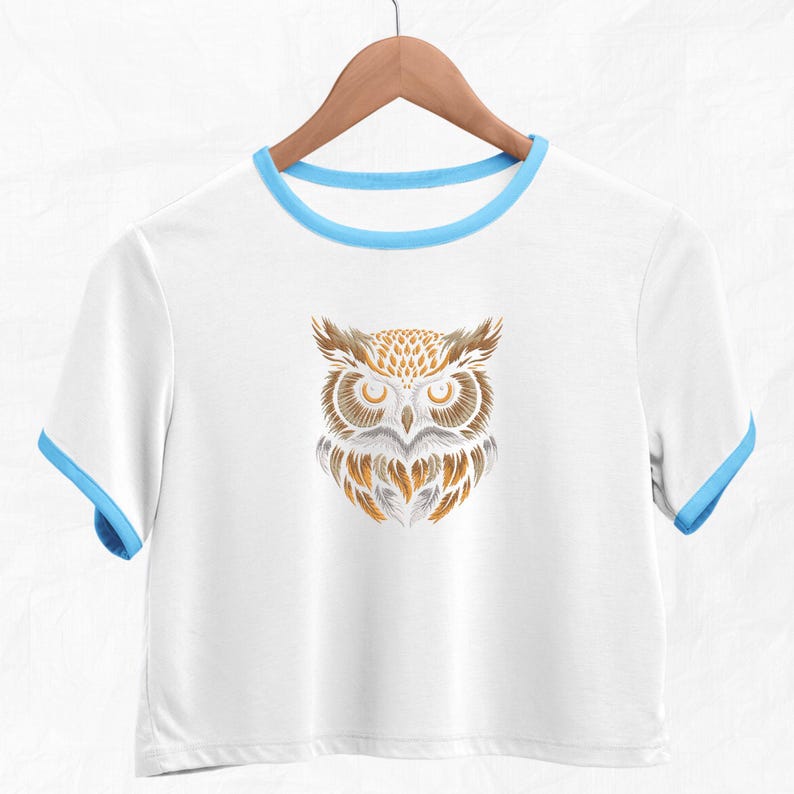 Op de afbeelding: Wit gecropped T-shirt met een blauwe rand rond de halslijn en mouwen. Het shirt heeft een afbeelding van een gestileerde uil met bruine en gouden veren.