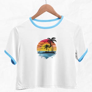 K&ouml;nnte beinhalten: Wei&szlig;es, bauchfreies T-Shirt mit blauem Saum am Ausschnitt. Das Shirt zeigt ein Motiv mit Palmen und Sonnenuntergang.