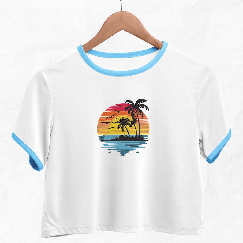 K&ouml;nnte beinhalten: Wei&szlig;es, bauchfreies T-Shirt mit blauem Saum am Ausschnitt. Das Shirt zeigt ein Motiv mit Palmen und Sonnenuntergang.