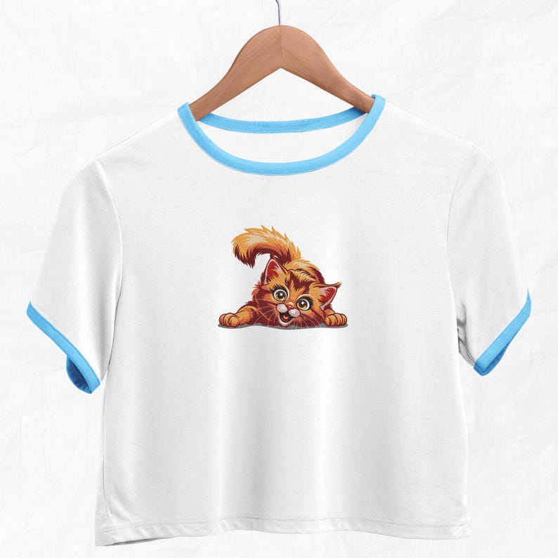 Op de afbeelding: Witte crop top met een lichtblauwe rand en een cartoonafbeelding van een oranje kat. De kat heeft een speelse pose, met uitgestrekte poten en een verraste uitdrukking. Het shirt hangt aan een houten hanger.