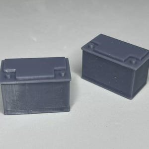 May include: Two gray rectangular car batteries. The batteries are made of plastic and have a textured surface.