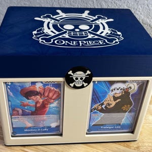 May include: A white and navy blue box featuring the One Piece logo and a skull and crossbones emblem. The box holds two trading cards, one featuring Monkey D. Luffy and the other Trafalgar Law.