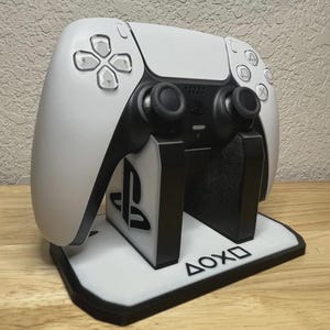 May include: A white and black gaming controller resting on a matching stand. The stand features the PlayStation logo and the symbols for the controller buttons. The controller has black joysticks and buttons.