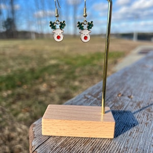 May include: A pair of gold-tone reindeer earrings with red noses and green antlers. The earrings are hanging from a wooden earring stand.