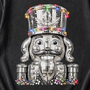 May include: A charcoal gray short-sleeve t-shirt featuring a graphic of a nutcracker made of silver mirrored disco ball tiles. The nutcracker wears a tall hat adorned with small, colorful glowing lights, and similar lights are at the base.