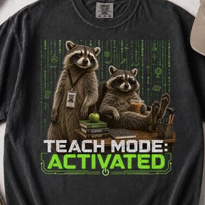 May include: A charcoal gray t-shirt featuring a graphic of two raccoons at a desk with books, an apple, and a drink. The background has green digital code patterns. The text reads TEACH MODE: ACTIVATED in white and green block lettering.