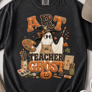May include: A charcoal grey t-shirt featuring a graphic of a ghost wearing a witch hat and glasses, holding a paintbrush and palette. The text reads ART TEACHER GHOST, surrounded by art supplies, autumn leaves, and a jack-o'-lantern pumpkin.