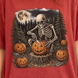 May include: A brick red short-sleeve t-shirt featuring a graphic print of a skeleton carving a jack-o'-lantern on a tree stump. The scene includes a full moon, a dark forest background, and two additional carved pumpkins on the ground.