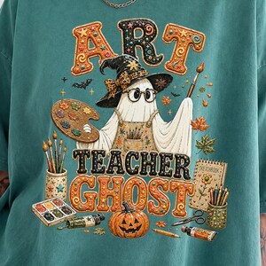 May include: A teal short-sleeve t-shirt featuring a graphic of a ghost wearing a witch hat and glasses, holding a paintbrush and palette. The text reads ART TEACHER GHOST, surrounded by art supplies, a pumpkin, and autumn leaves in orange and brown.