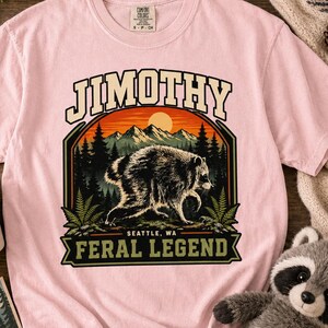 May include: A light pink short-sleeve t-shirt featuring a graphic of a raccoon in a forest with mountains and a sun. The text reads JIMOTHY at the top and FERAL LEGEND SEATTLE, WA at the bottom. The shirt has a crew neckline and a standard fit.