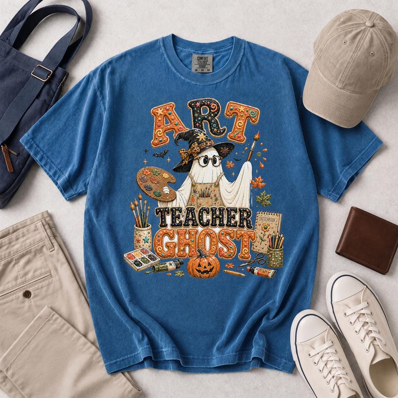 May include: A blue t-shirt featuring a graphic of a ghost wearing a witch hat and glasses, holding a paintbrush and palette. The text reads ART TEACHER GHOST. The design includes a pumpkin, paintbrushes, a sketchbook, and paint supplies.