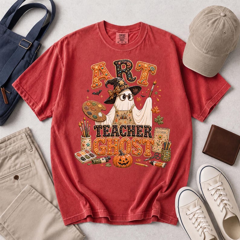 May include: A red short-sleeved t-shirt featuring a graphic of a ghost wearing a witch hat and glasses, holding a paintbrush. The text reads ART TEACHER GHOST. The design includes a paint palette, brushes, a pumpkin, and art supplies.