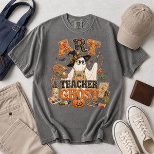 May include: A charcoal gray t-shirt featuring a graphic of a ghost wearing a witch hat, glasses, and an apron. The ghost holds a paintbrush and palette. Text reads ART TEACHER GHOST, surrounded by art supplies, a pumpkin, and autumn leaves.