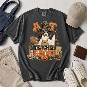 May include: A charcoal gray t-shirt featuring a graphic of a ghost wearing a witch hat, glasses, and an apron. The ghost holds a paintbrush and palette. The text reads ART TEACHER GHOST, surrounded by art supplies, a pumpkin, and autumn leaves.
