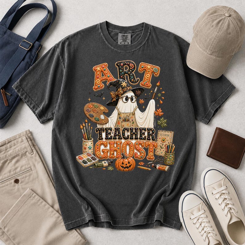 May include: A charcoal gray t-shirt featuring a graphic of a ghost wearing a witch hat, glasses, and an apron. The ghost holds a paintbrush and palette. The text reads ART TEACHER GHOST, surrounded by art supplies, a pumpkin, and autumn leaves.