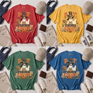 May include: A collection of four short-sleeve t-shirts in red, yellow, green, and blue. Each shirt features a graphic of a ghost wearing a witch hat, holding a paintbrush and palette, with the text ART TEACHER GHOST and various art supplies.
