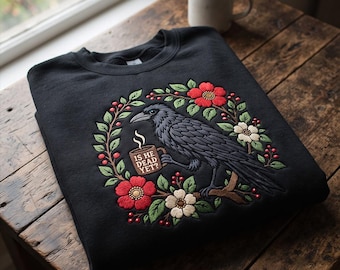 May include: A black sweatshirt featuring an embroidered design of a black crow perched on a branch, holding a brown mug with the text IS HE DEAD YET? surrounded by a wreath of red and white flowers, green leaves, and small red berries.