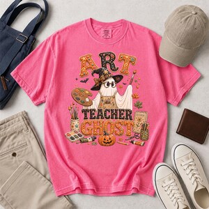 May include: A bright pink t-shirt featuring a graphic of a ghost wearing a witch hat and glasses, holding a paintbrush and palette. The text reads ART TEACHER GHOST, surrounded by art supplies, autumn leaves, and a pumpkin on a light background.