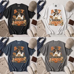 May include: Four short-sleeve t-shirts in black, white, blue, and gray, each featuring a graphic of a ghost wearing a witch hat and holding a paintbrush. The design includes a paint palette, pumpkin, and the text ART TEACHER GHOST.