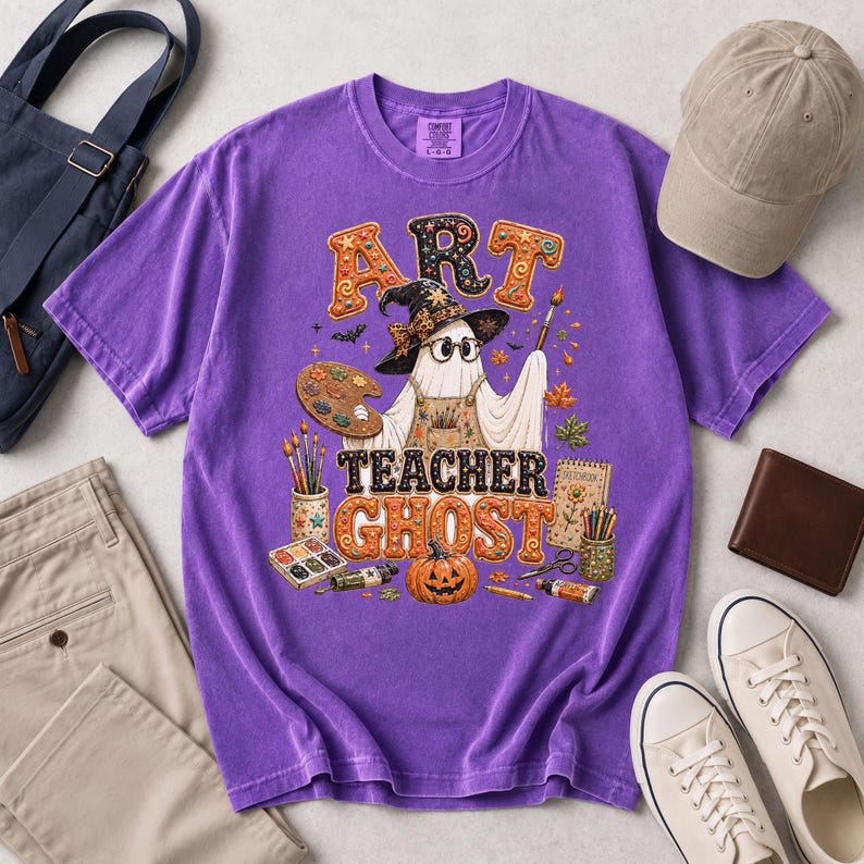 May include: A purple short-sleeve t-shirt featuring a graphic of a ghost wearing a witch hat and glasses, holding a paintbrush. The text reads ART TEACHER GHOST. The design includes a paint palette, a pumpkin, brushes, and a sketchbook.